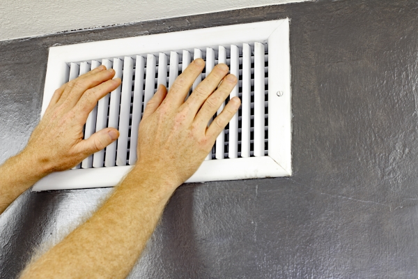 A pair of adult male hands feeling the flow of air coming out of an air vent on a wall near a ceiling in a residential home.