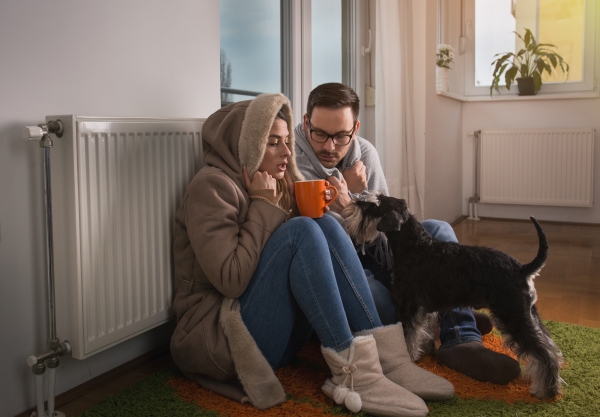 Young couple in home wearing a jacket and covered with blanket sitting on floor beside radiator with dog and trying to warm up